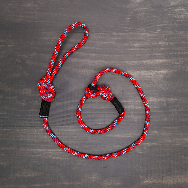 The Maple Slip Lead, made from durable rock climbing rope in red, white, and black colors, is laid out flat on a gray wooden surface. It features a knot at one end and a sliding loop at the other, making it ideal for training sessions and built to last.
